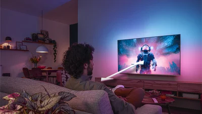 Man zit op een bank en kijkt naar een Philips Ambilight-tv met een kleurrijke astronautscène, waarbij een pijl de optimale kijkafstand aangeeft