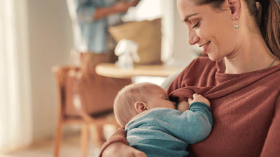 A mother wearing a pink shirt, sitting on a chair, breastfeeding her baby.