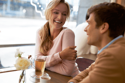 Une femme blonde souriante est assise à une table avec un jeune homme, face à une tasse de thé et un café