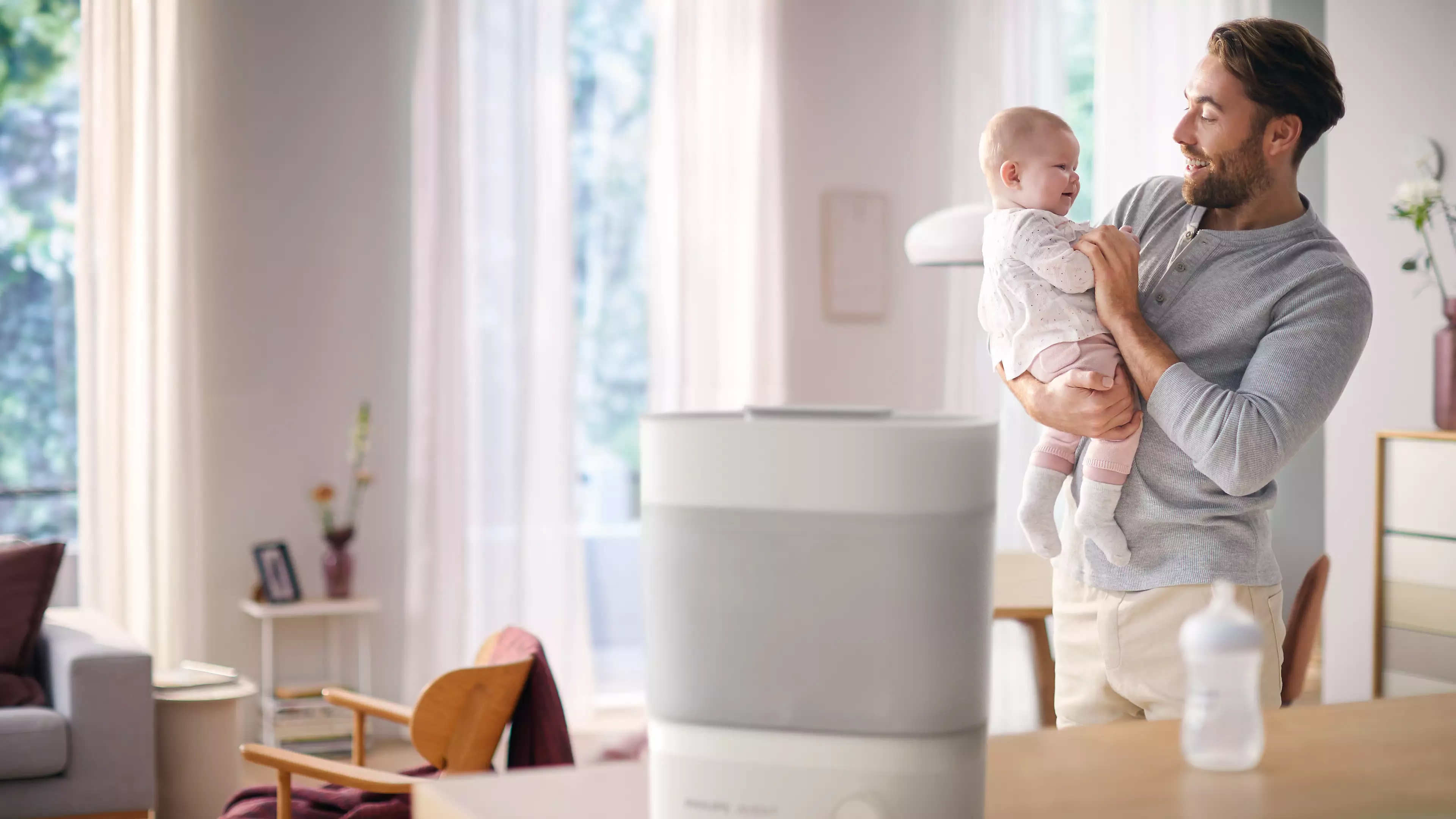 Sterylizator butelek Philips Avent i tata trzymający dziecko na rękach w tle