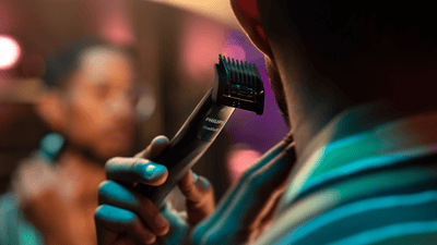 Product close-up of a Philips OneBlade and body comb trimming sideburns of a man looking into a mirror.
