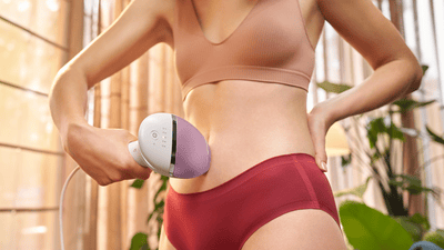 A woman in nude and burgundy underwear is using an IPL device to remove hair on her stomach.