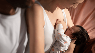 A woman and a man just out of view look down at their young infant, while the woman breastfeeds on a soft surface.