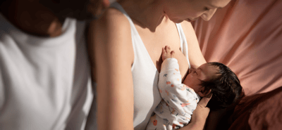 A woman and a man just out of view look down at their young infant, while the woman breastfeeds on a soft surface.