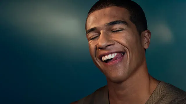A young man smiles as he licks his white teeth against a dark gradient background.