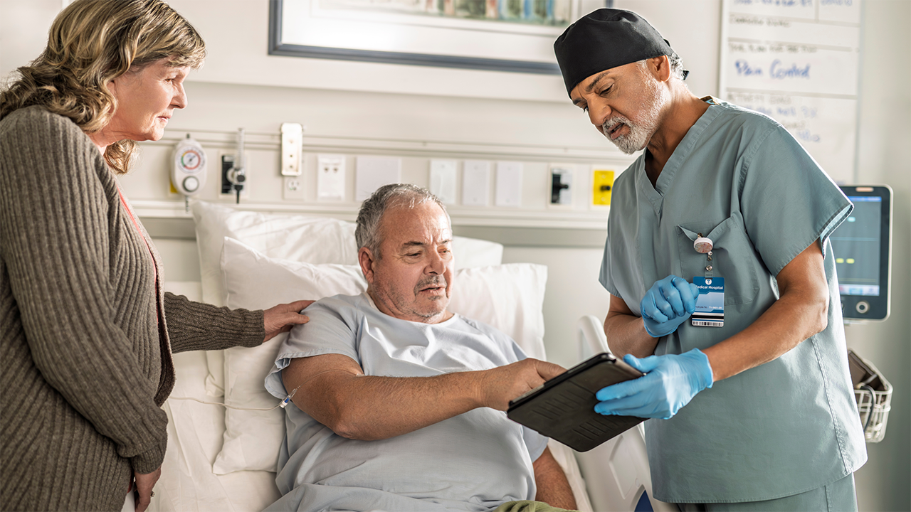 A patient in a hospital gown is seated on an examination bed. Two healthcare providers are present — one appears to hold a clipboard or document. The calm collaborative scene conveys reassurance and clinical professionalism, contextualising the endoscopy section.