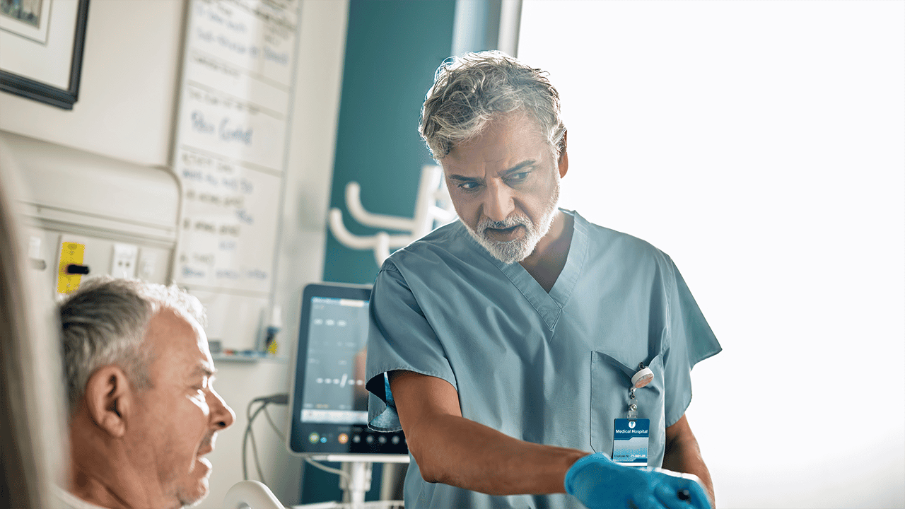 Male clinician in surgical scrubs attending to a patient lying in a hospital bed A male clinician in teal scrubs and blue gloves attends to a patient in a hospital room. A clinical whiteboard and equipment are visible in the background, contextualising the endoscopy and biopsy diagnostic procedure described alongside.