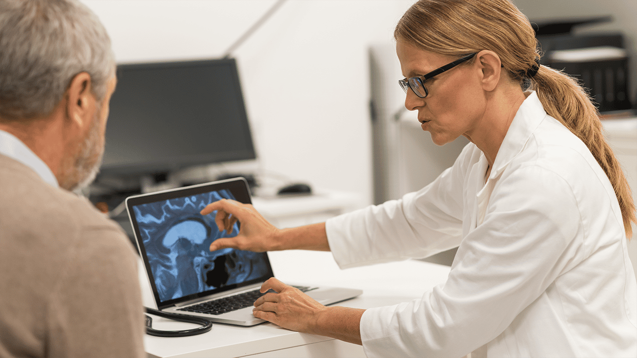A doctor in a white coat sits alongside a patient, both reviewing what appears to be a medical scan on a screen or tablet. Reflects collaborative review of imaging results and contextualises MRI and ultrasound as patient-accessible monitoring tools.