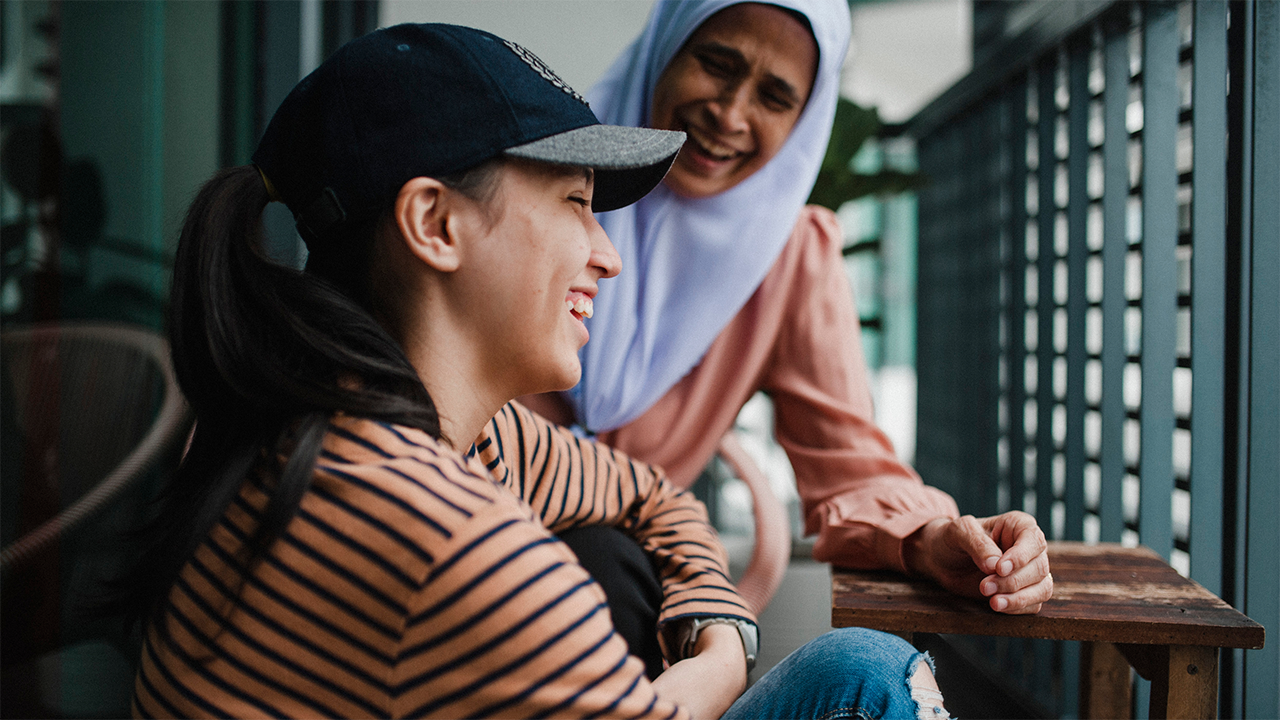 A young woman in a striped top and baseball cap smiles up at an older woman in a lavender hijab on an outdoor terrace, conveying the warmth and connection of a peer support community.