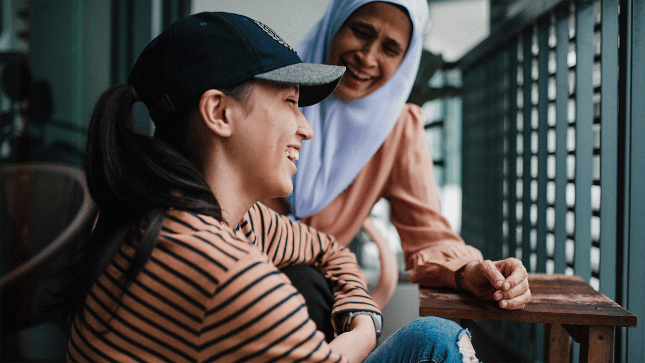 A young woman in a striped top and baseball cap smiles up at an older woman in a lavender hijab on an outdoor terrace, conveying the warmth and connection of a peer support community.