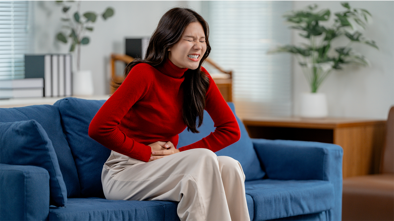 A woman in a red top sits on a blue sofa with one hand on her abdomen and her gaze downward. The image conveys physical discomfort, representing the flares and triggers theme of this FAQ section.