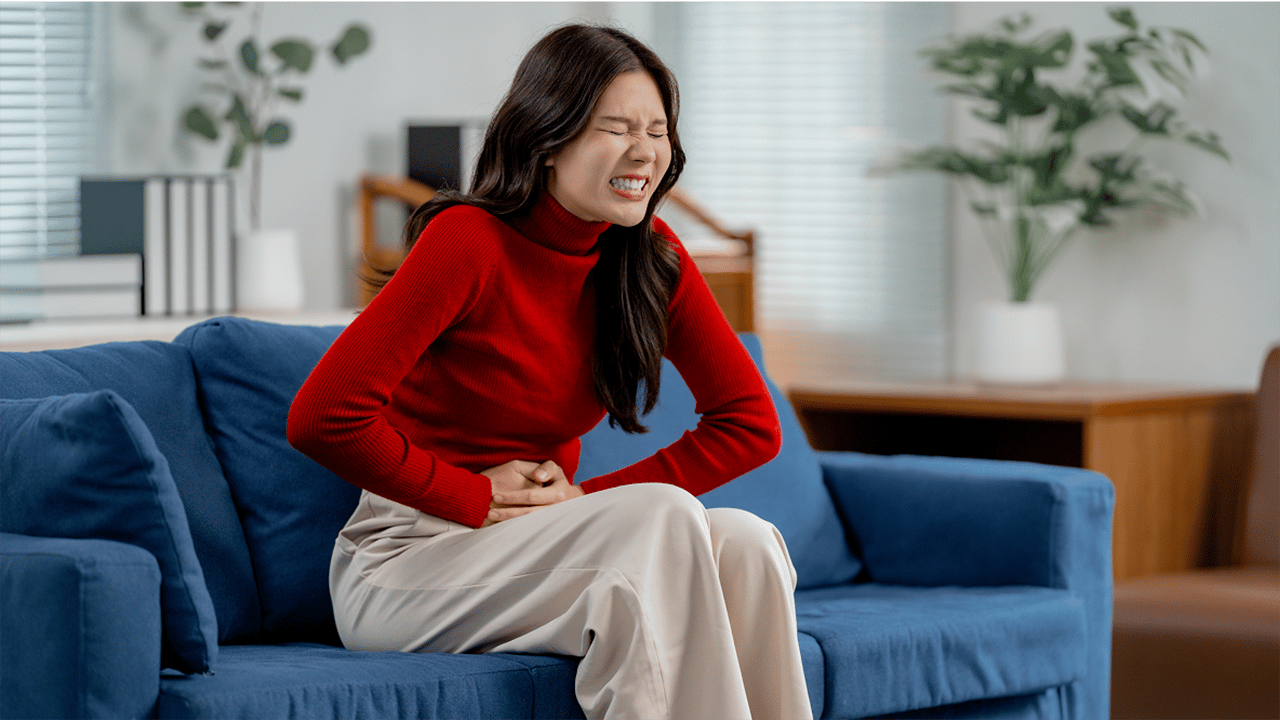 A woman in a red top sits on a blue sofa with one hand on her abdomen and her gaze downward. The image conveys physical discomfort, representing the flares and triggers theme of this FAQ section.