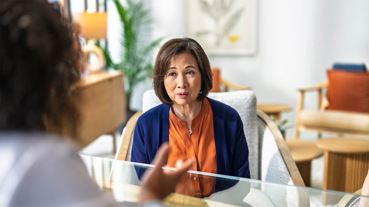 An Asian woman in warm-toned attire appears confident and animated in a relaxed doctor's office. Her open, engaged expression conveys the empowerment and voice that comes from feeling heard and supported within a healthcare context.
