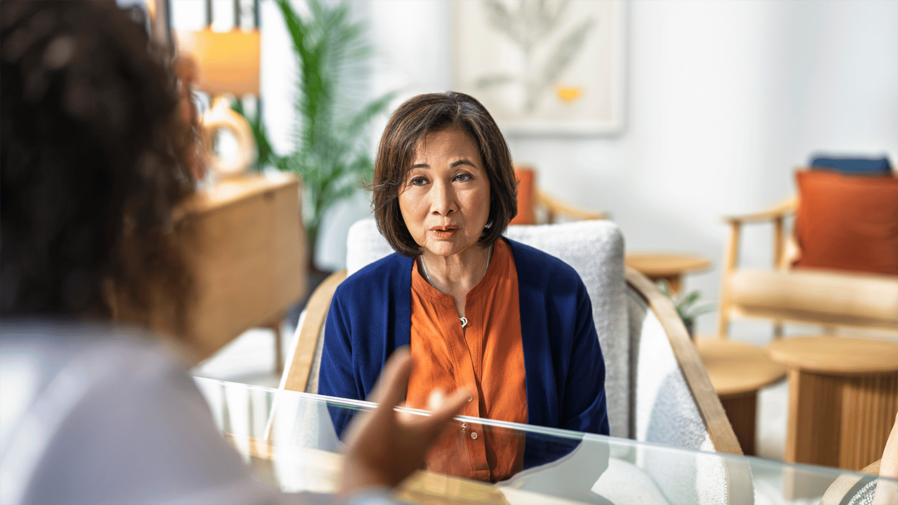 An Asian woman in warm-toned attire appears confident and animated in a relaxed doctor's office. Her open, engaged expression conveys the empowerment and voice that comes from feeling heard and supported within a healthcare context.