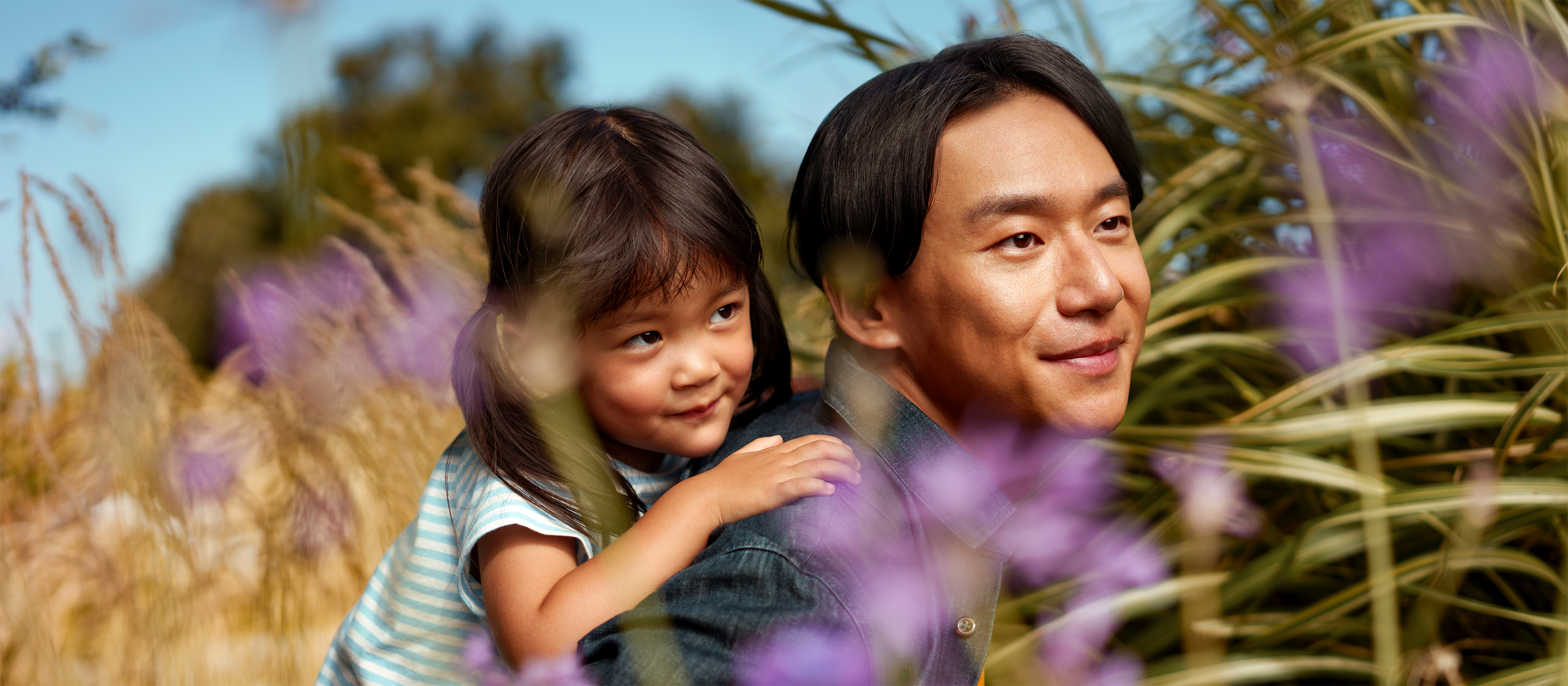 A smiling Asian man stands outdoors as a young girl hugs him from behind, both surrounded by purple wildflowers and tall grass beneath a blue sky. Warm lifestyle photography conveying family, joy, and everyday wellbeing.