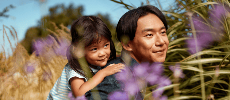 Father and young daughter outdoors surrounded by purple wildflowers A smiling Asian man stands outdoors as a young girl hugs him from behind, both surrounded by purple wildflowers and tall grass beneath a blue sky. Warm lifestyle photography conveying family, joy, and everyday wellbeing.