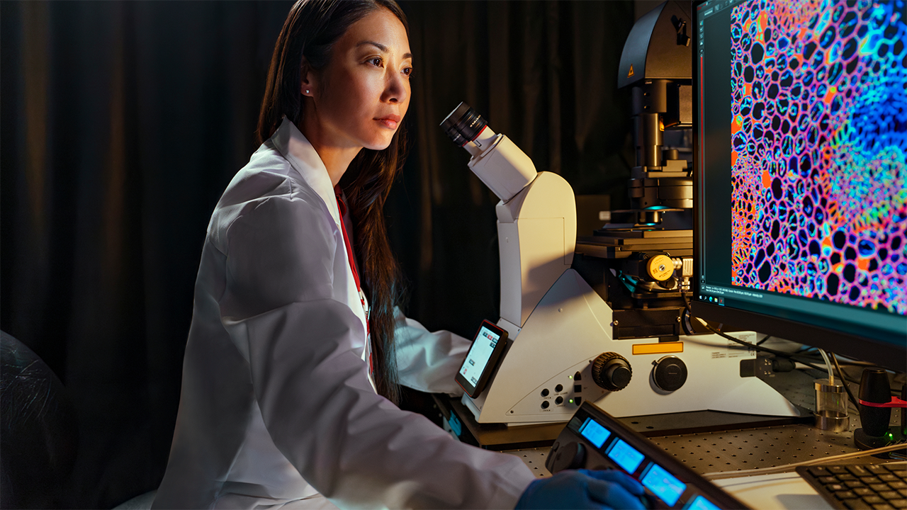 A female researcher in a white coat works at an advanced microscope station in a dark laboratory, with vivid microscopy images glowing on screens behind her. Evokes high-technology diagnostics, supporting the MRI and CT scans section.