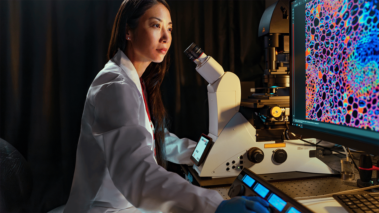Female researcher in white lab coat at a microscope workstation with colourful imaging screens A female researcher in a white coat works at an advanced microscope station in a dark laboratory, with vivid microscopy images glowing on screens behind her. Evokes high-technology diagnostics, supporting the MRI and CT scans section.