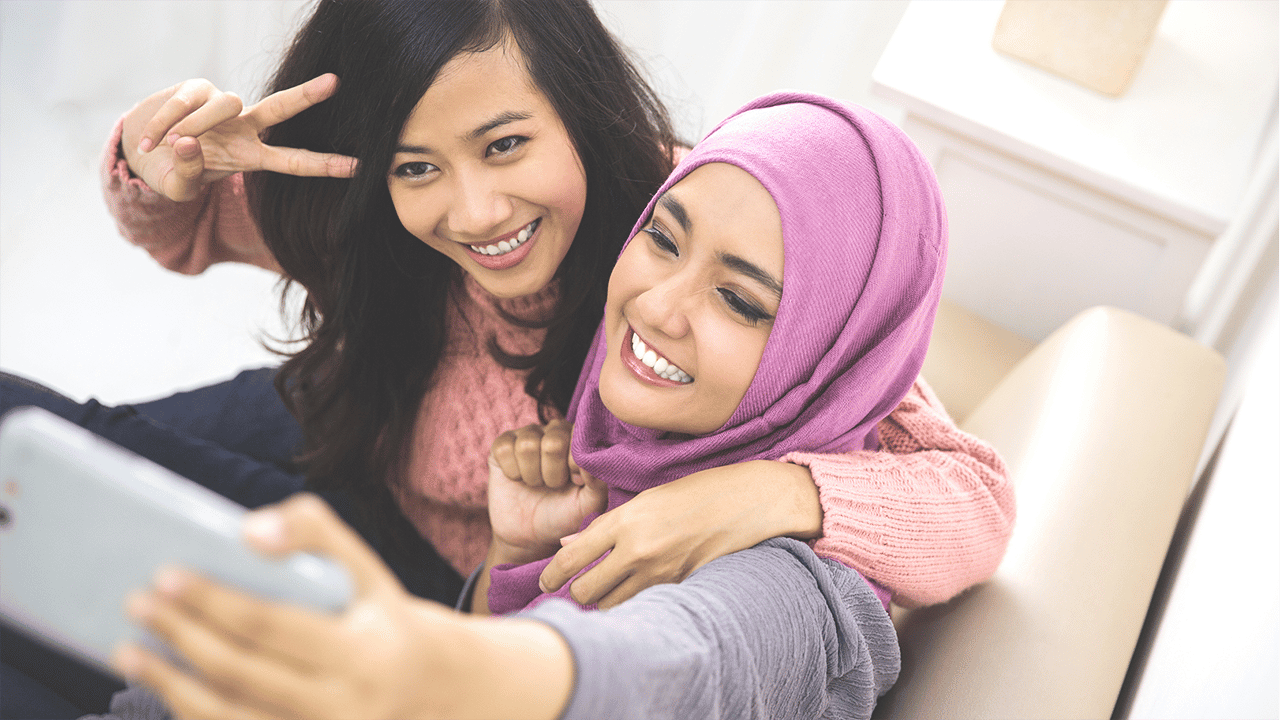 Two women share a moment looking at a phone screen together. One wears a pink hijab. Both express visible happiness or engagement. The casual upbeat scene accompanies content about endoscopic remission — the point where a gastroenterologist can confirm tissue-level healing.