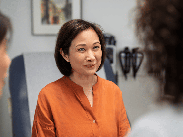 ibd-healthcare-journey An Asian woman in an orange top sits in a clinical consultation setting, looking toward a healthcare professional visible in the background. The image represents a patient actively participating in a shared healthcare discussion.