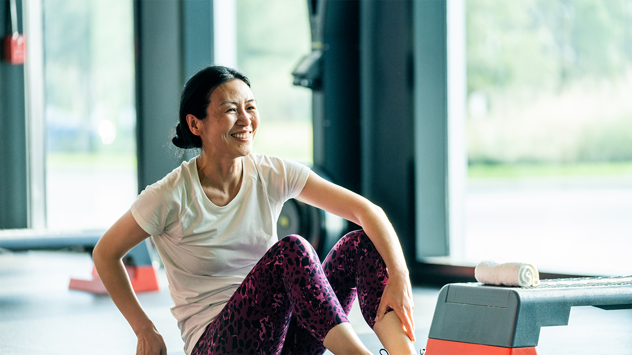 Asian woman is smiling while exercising in a bright, airy gym with lots of natural light. Suggests the confident that comes with shared decision making.