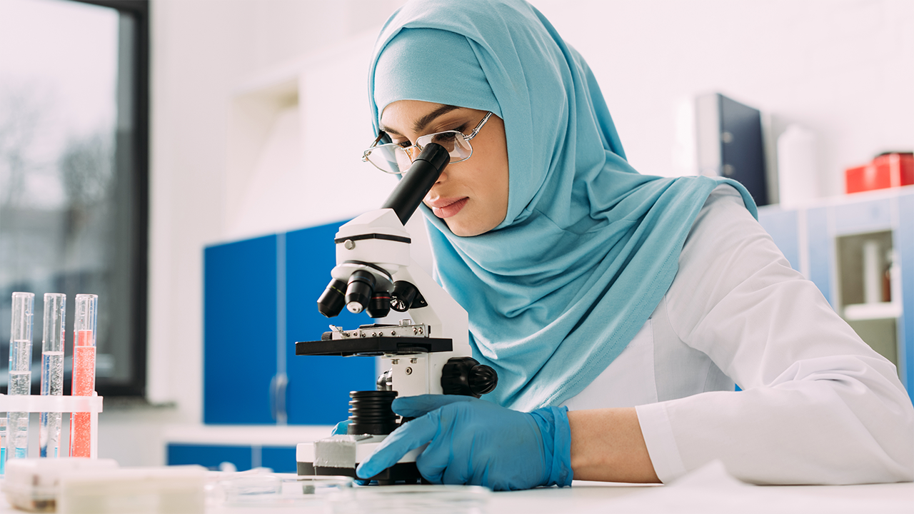 A female laboratory technician in a light blue hijab and protective gloves uses a microscope in a well-lit clinical lab. Colourful test tubes in the background illustrate laboratory analysis as described in the blood and stool test diagnosis section.