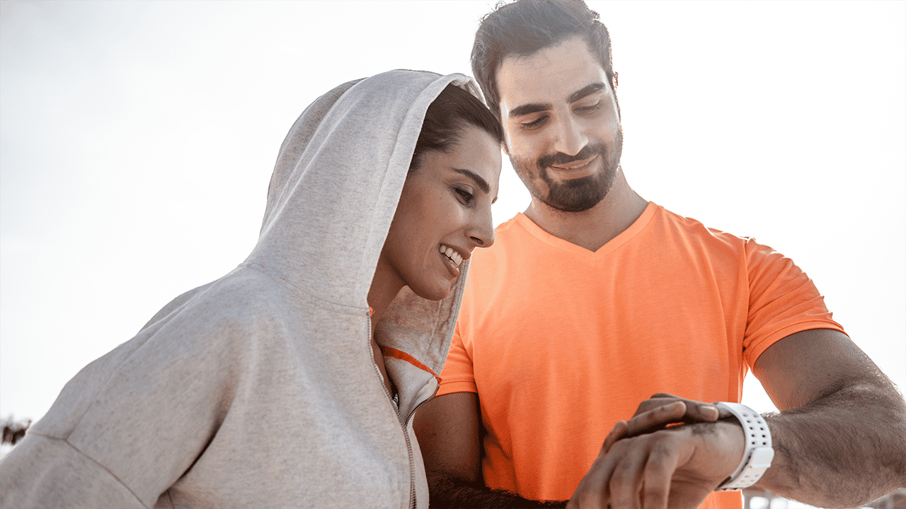 A smiling couple sharing a light-hearted moment while exercising together, reflecting on their progress via a smartwatch. Positioned alongside the 'Set and measure your own goals' tip about planning achievable daily and long-term goals.
