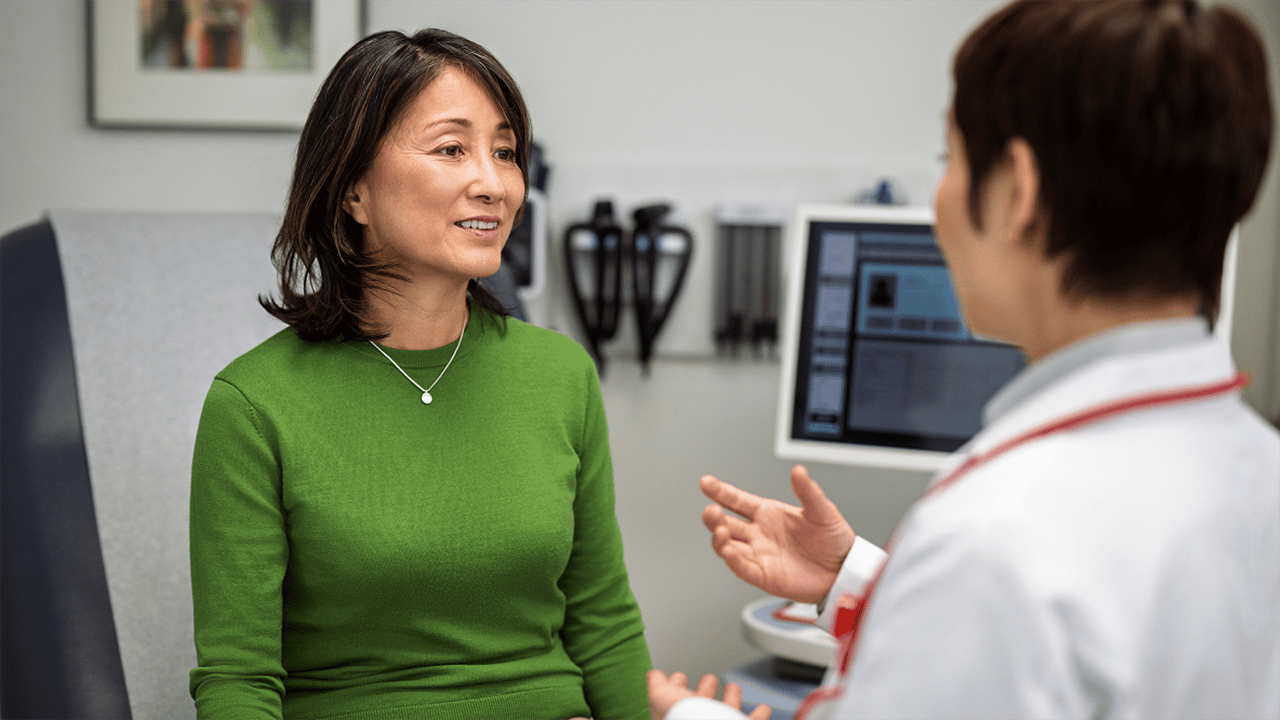 A person wearing a dark green top sits opposite a healthcare professional in a white coat in what appears to be a clinical consultation room, representing a doctor-patient discussion about Crohn's disease.