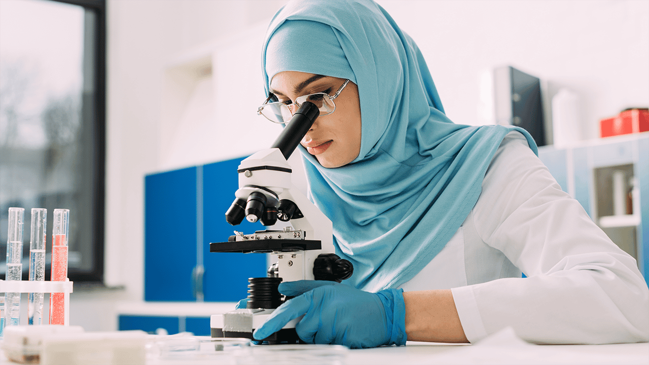 A female laboratory technician in a light blue hijab and protective gloves uses a microscope in a well-lit clinical lab. Colourful test tubes in the background illustrate laboratory analysis as described in the blood and stool test diagnosis section.