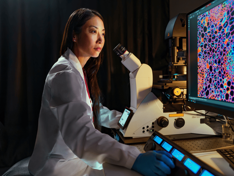 envision An asian woman in a white lab coat looks into a microscope in a darkened biomedical research laboratory. Behind her, large computer monitors display vivid cellular imagery, suggesting active scientific research and discovery.