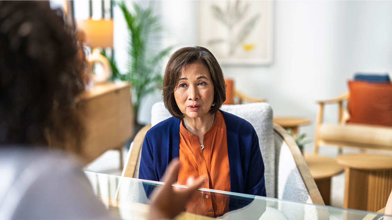 An Asian woman in warm-toned attire appears confident and animated in a relaxed doctor's office. Her open, engaged expression conveys the empowerment and voice that comes from feeling heard and supported within flare-ups and day to day management of her condition.