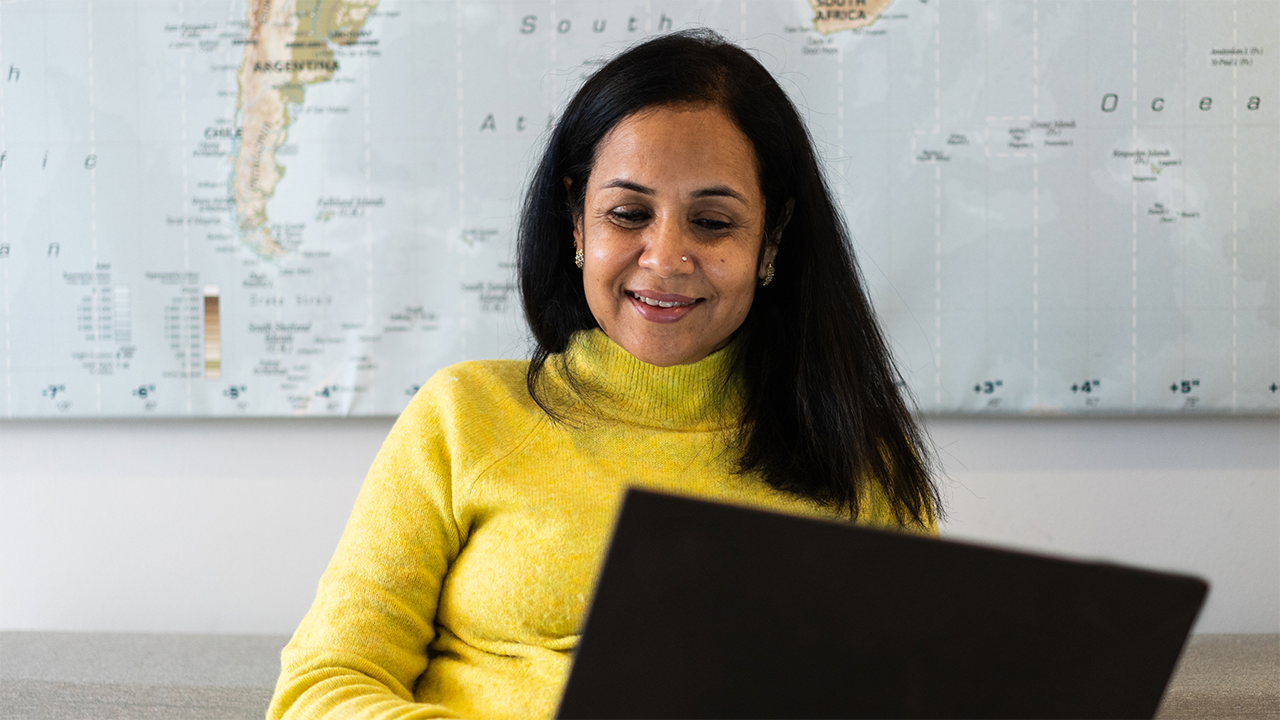 A South Asian woman in a bright yellow turtleneck smiles warmly while using a laptop, with a world map on the wall behind her, conveying the habit of self-tracking and positive engagement with health management.