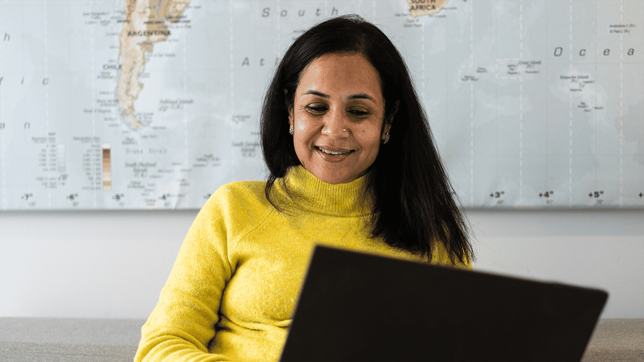 A South Asian woman in a bright yellow turtleneck smiles warmly while using a laptop, with a world map on the wall behind her, conveying the habit of self-tracking and positive engagement with health management.