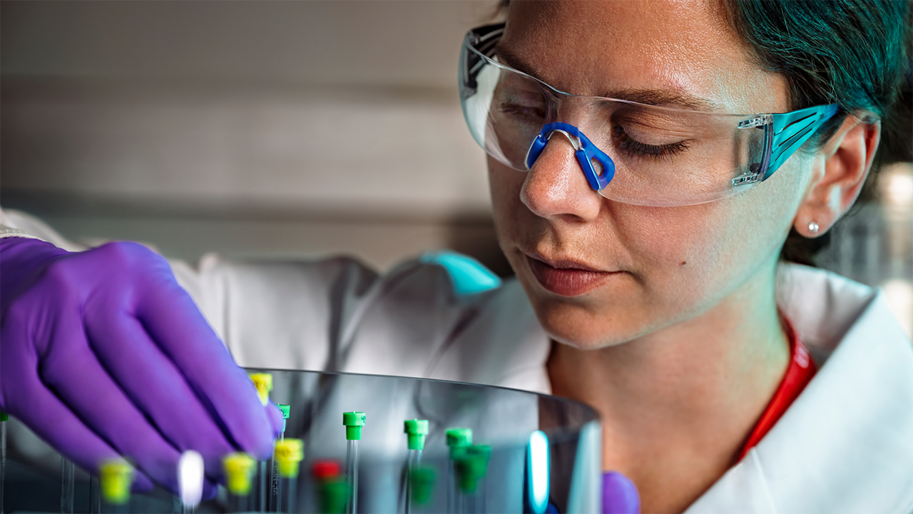 A laboratory worker in blue nitrile gloves and protective safety goggles holds glass test tubes with coloured liquid samples. The clinical lab setting reinforces the precision of blood-based biomarker testing — contextualising the C-reactive protein (CRP) measurement used to monitor IBD activity.