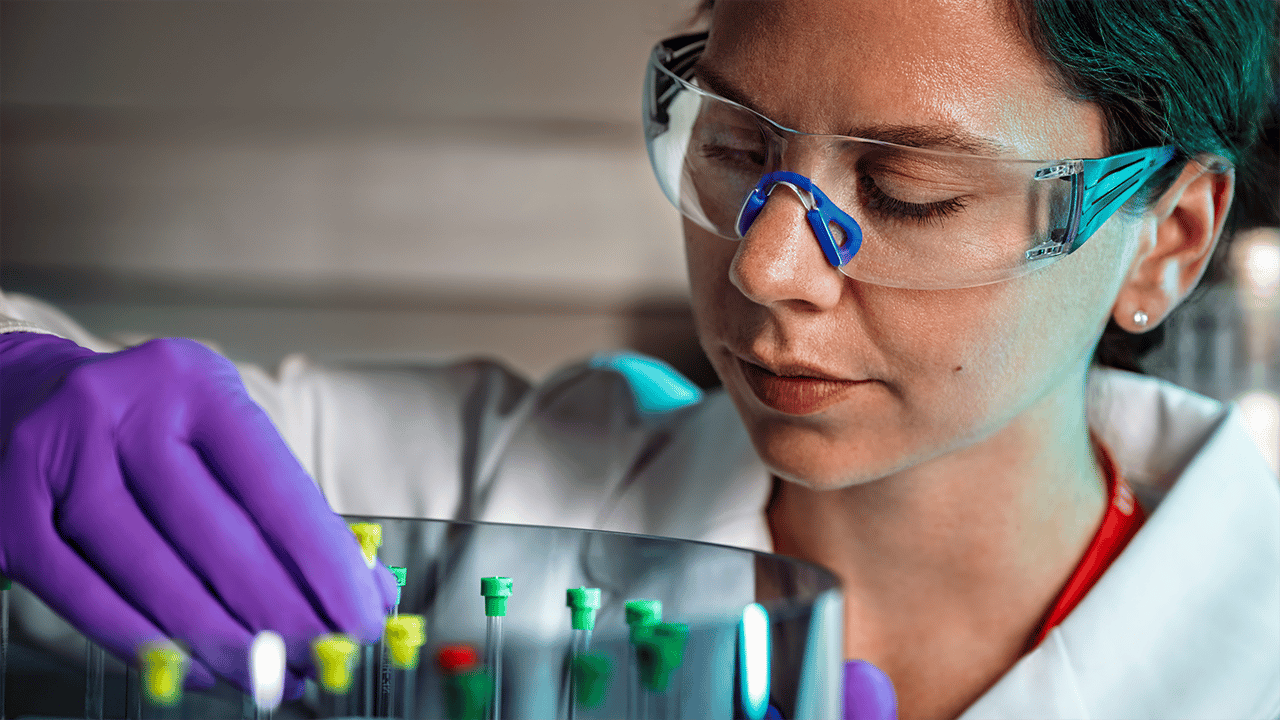 A laboratory worker in blue nitrile gloves and protective safety goggles holds glass test tubes with coloured liquid samples. The clinical lab setting reinforces the precision of blood-based biomarker testing — contextualising the C-reactive protein (CRP) measurement used to monitor IBD activity.