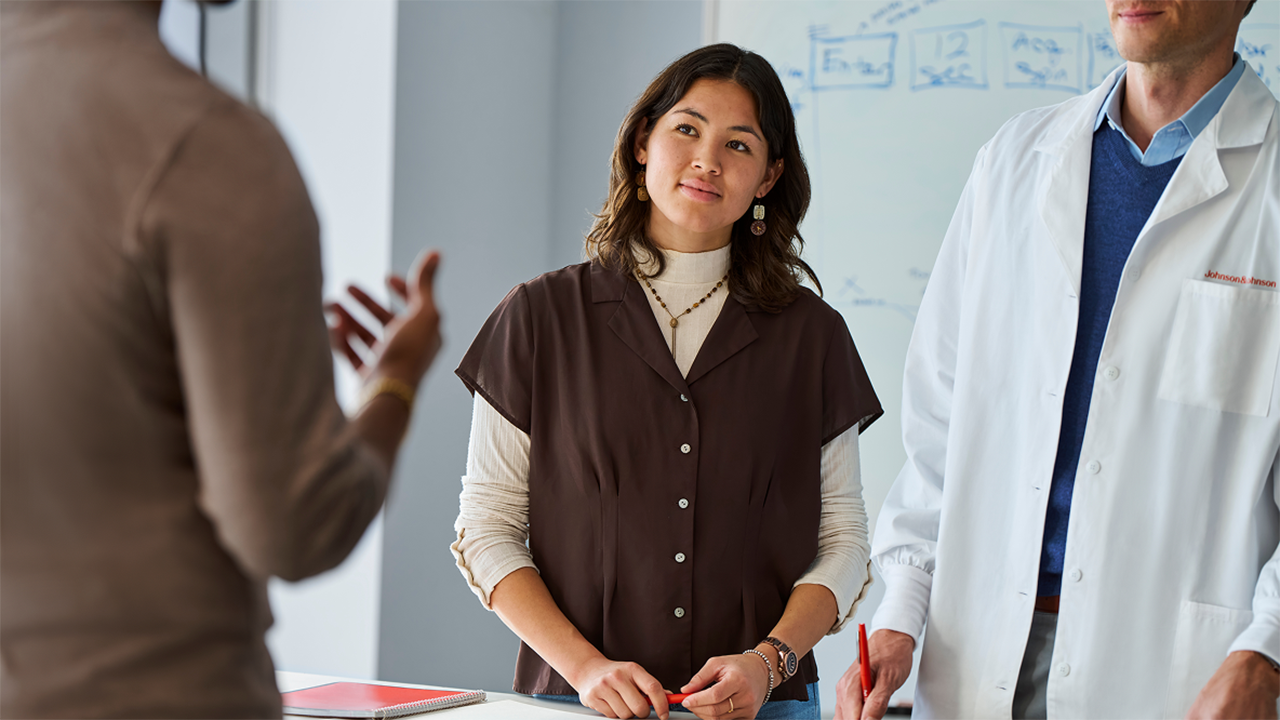 A asian woman photographed in a professional or workplace meeting setting, engaged in discussion. The image conveys active participation in work life alongside a chronic condition.