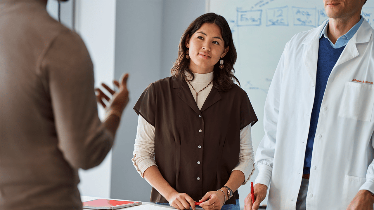 A asian woman photographed in a professional or workplace meeting setting, engaged in discussion. The image conveys active participation in work life alongside a chronic condition.