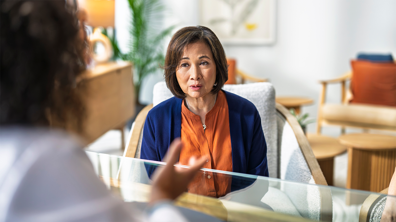 A middle-aged woman in an orange top sits across from a healthcare professional in a warm, plant-filled consultation room. The approachable naturalistic setting illustrates the medical history stage of the ulcerative colitis diagnostic journey.