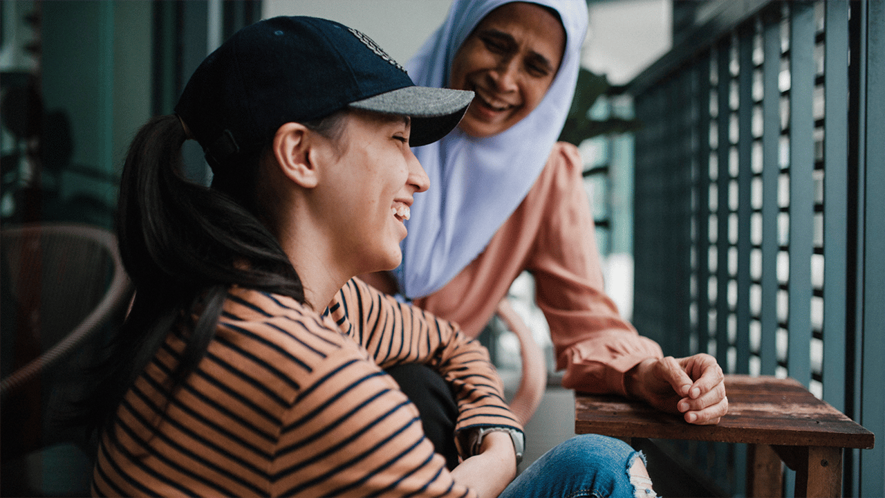A young woman in a striped top and baseball cap smiles up at an older woman in a lavender hijab on an outdoor terrace, conveying the warmth of a long term outlook.