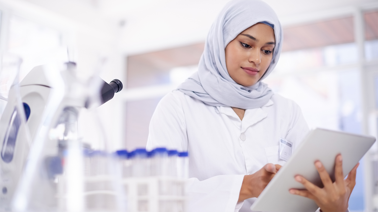 A woman wearing a white hijab and professional white lab coat stands in a clinical or laboratory environment, looking attentively at a digital tablet. The setting and attire suggest a healthcare professional context. Illustrates a non-invasive and technologically-aided approach to monitoring gut health.