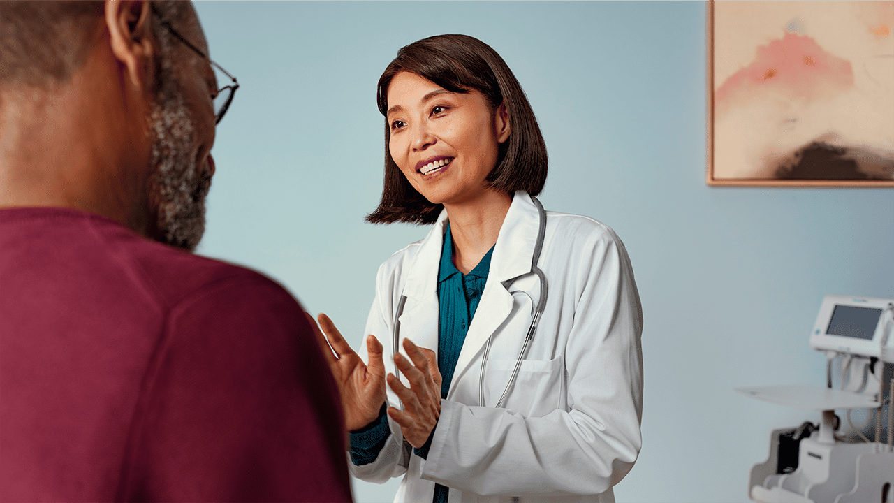 Female doctor in white coat and stethoscope smiling while speaking with a patient A female healthcare professional in a white coat and stethoscope smiles during a patient consultation. The positive, reassuring clinical exchange supports the physical examination step in the Crohn's disease diagnosis section.