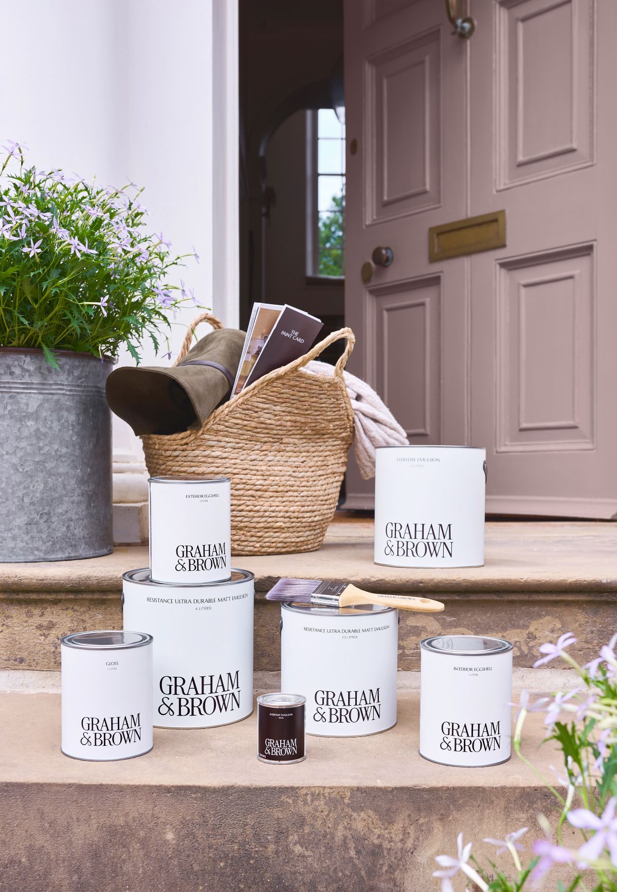Graham & Brown exterior paint tins arranged on the steps leading up to a partially open front door. A metal plant pot sits on the top step to the right, next to a wicker basket, framing a front door painted in Spiced Mulberry exterior eggshell.