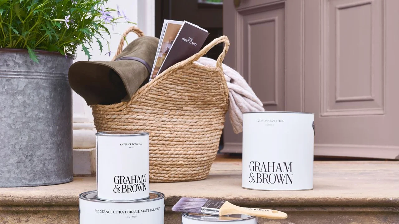 Graham & Brown exterior paint tins arranged on the steps leading up to a partially open front door. A metal plant pot sits on the top step to the right, next to a wicker basket, framing a front door painted in Spiced Mulberry exterior eggshell.