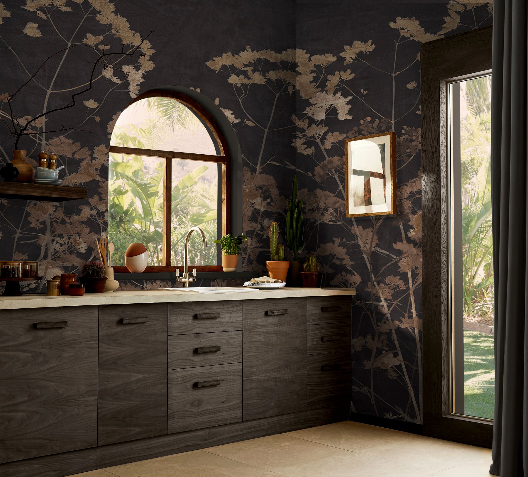 A modern kitchen featuring dark cabinets painted in Graham & Brown’s Fetish, paired with the Flower Press Chocolate mural, a light countertop, and large windows overlooking greenery, with potted plants and kitchen items arranged on the counter.