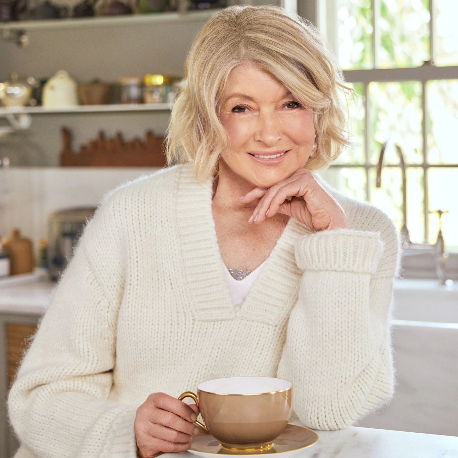 Martha Stewart sat at a table in a kitchen holding a cup and saucer wearing a white woollen jumper