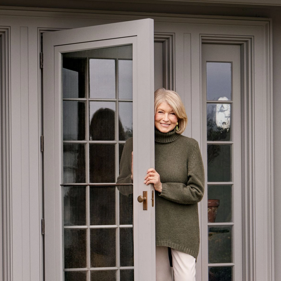 Martha Stewart at home in a grey wool jumper stood in the open doorway looking out