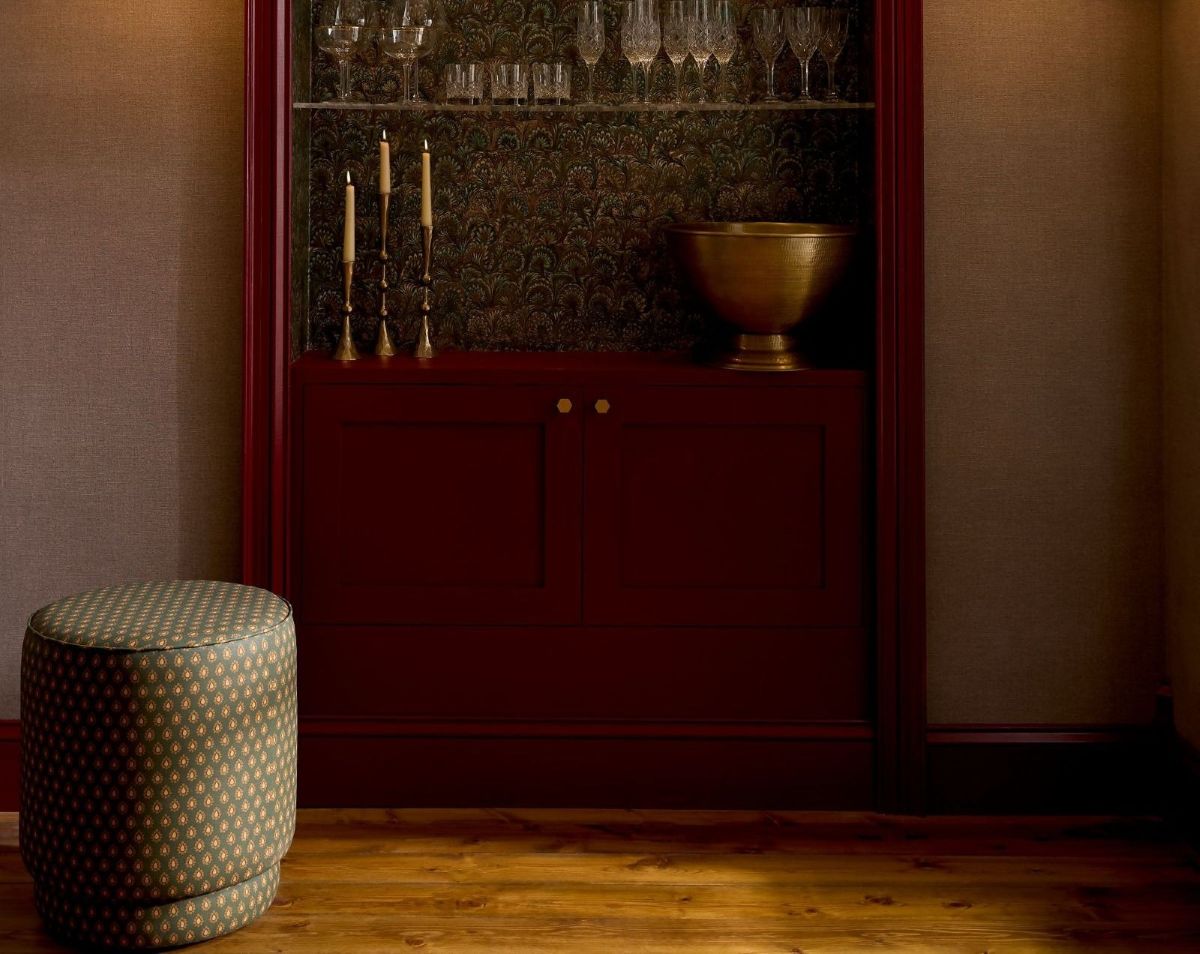 Truffle Peacock Wallpaper behind a bar cabinet which is painted in Roger Red Eggshell with Haptic Hazel on the walls and Dobby Forest Pouffe to the left of the bar area