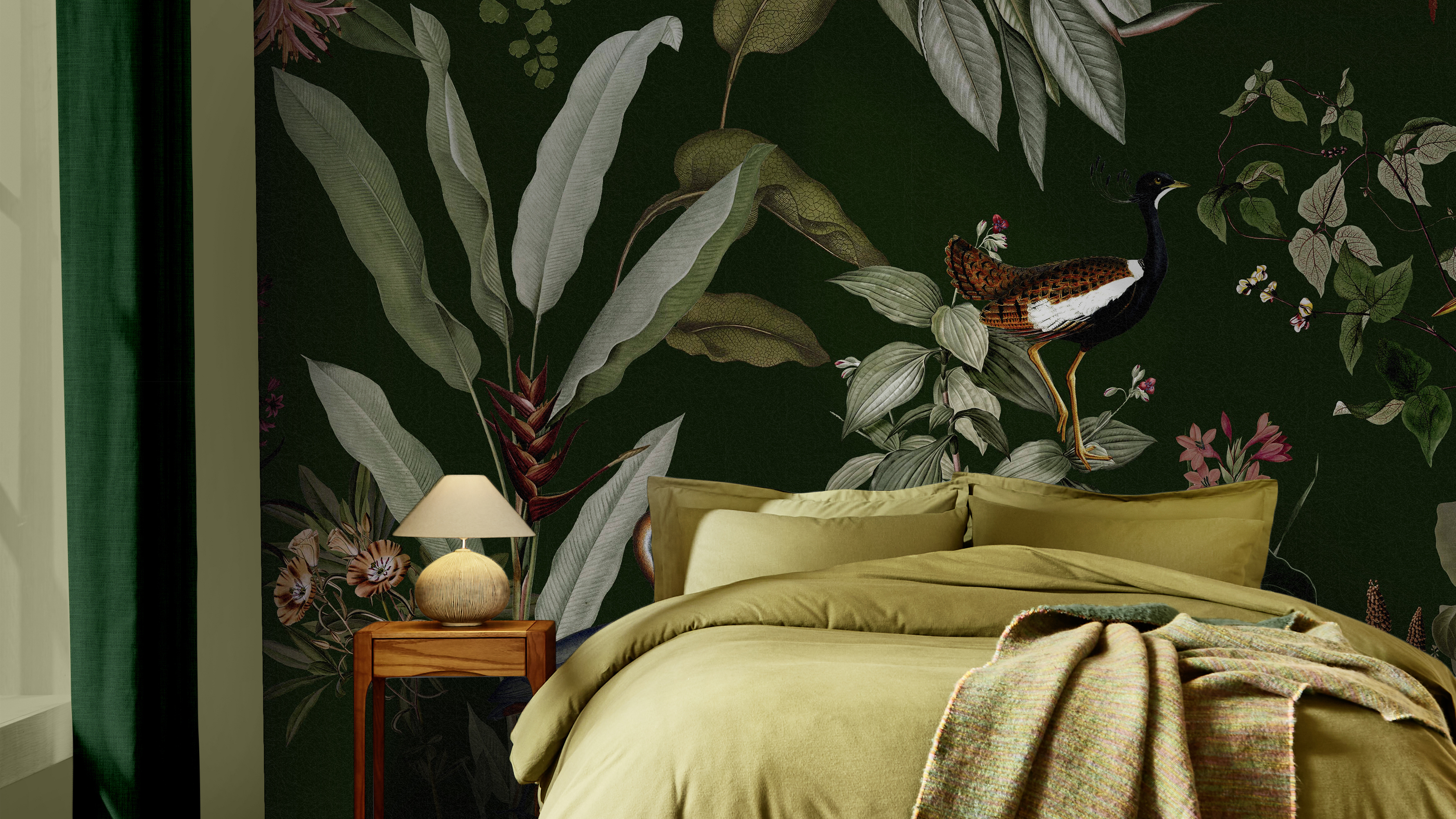 A bedroom with an olive‑green bedspread and matching pillows, a textured throw draped across the bed, and a wooden bedside table with a round lamp. The wall behind the bed features Graham & Brown's Glasshouse Bespoke Mural with large illustrated leaves, flowers, and a small bird perched among the foliage. A green curtain hangs to the left, creating a calm, nature‑inspired atmosphere.