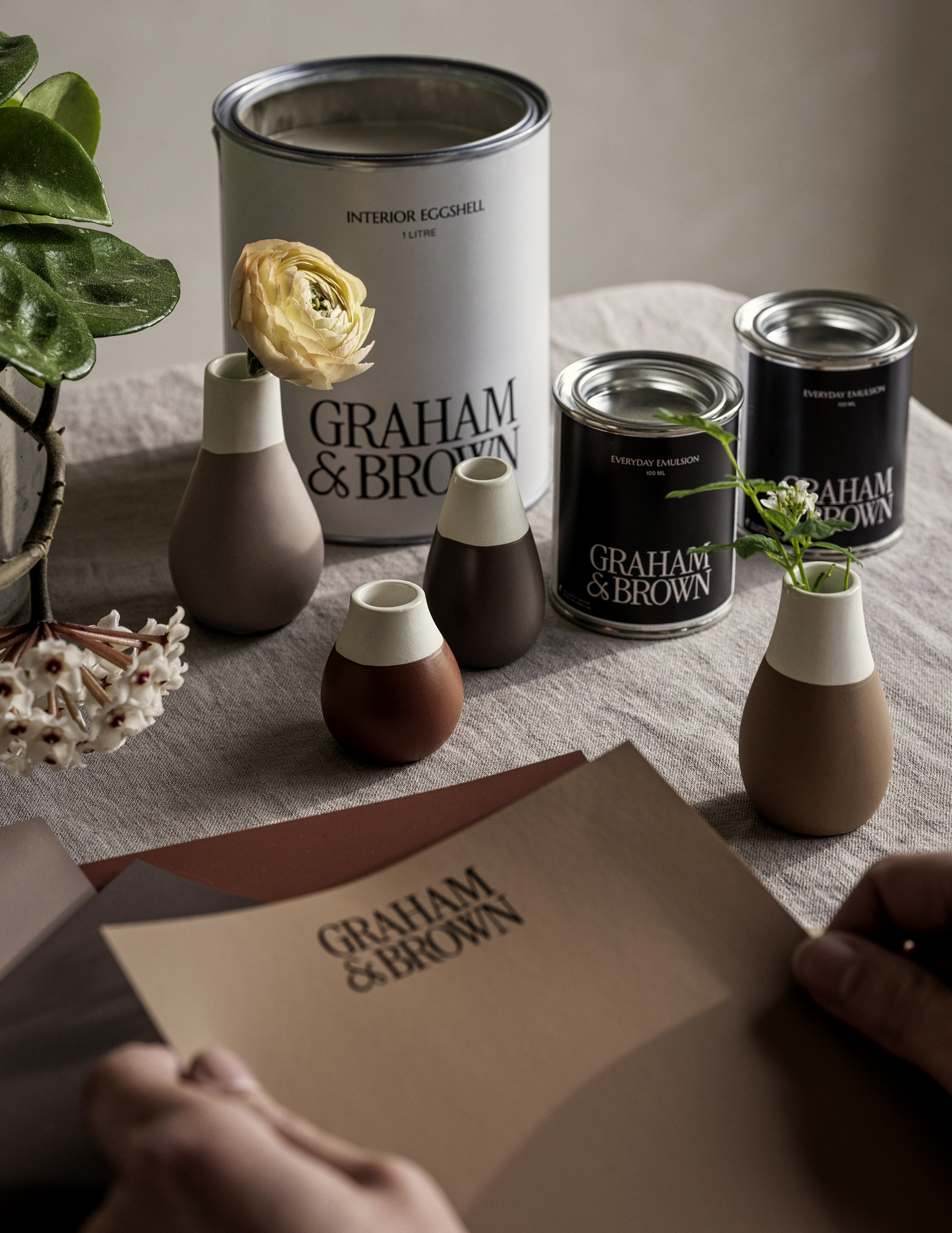Hands holding peel and stick paint samples of the create colour story on a dining room table with 3 paint tins and decor. 