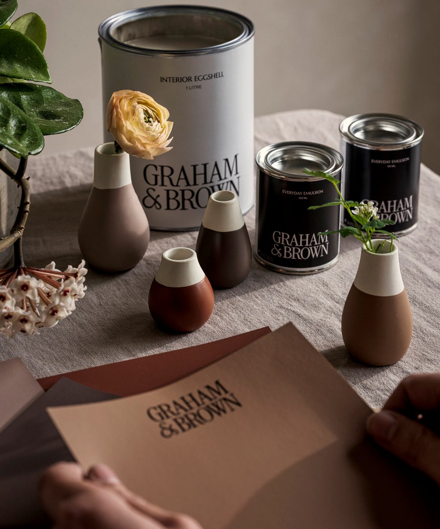 Hands holding peel and stick paint samples of the create colour story on a dining room table with 3 paint tins and decor.
